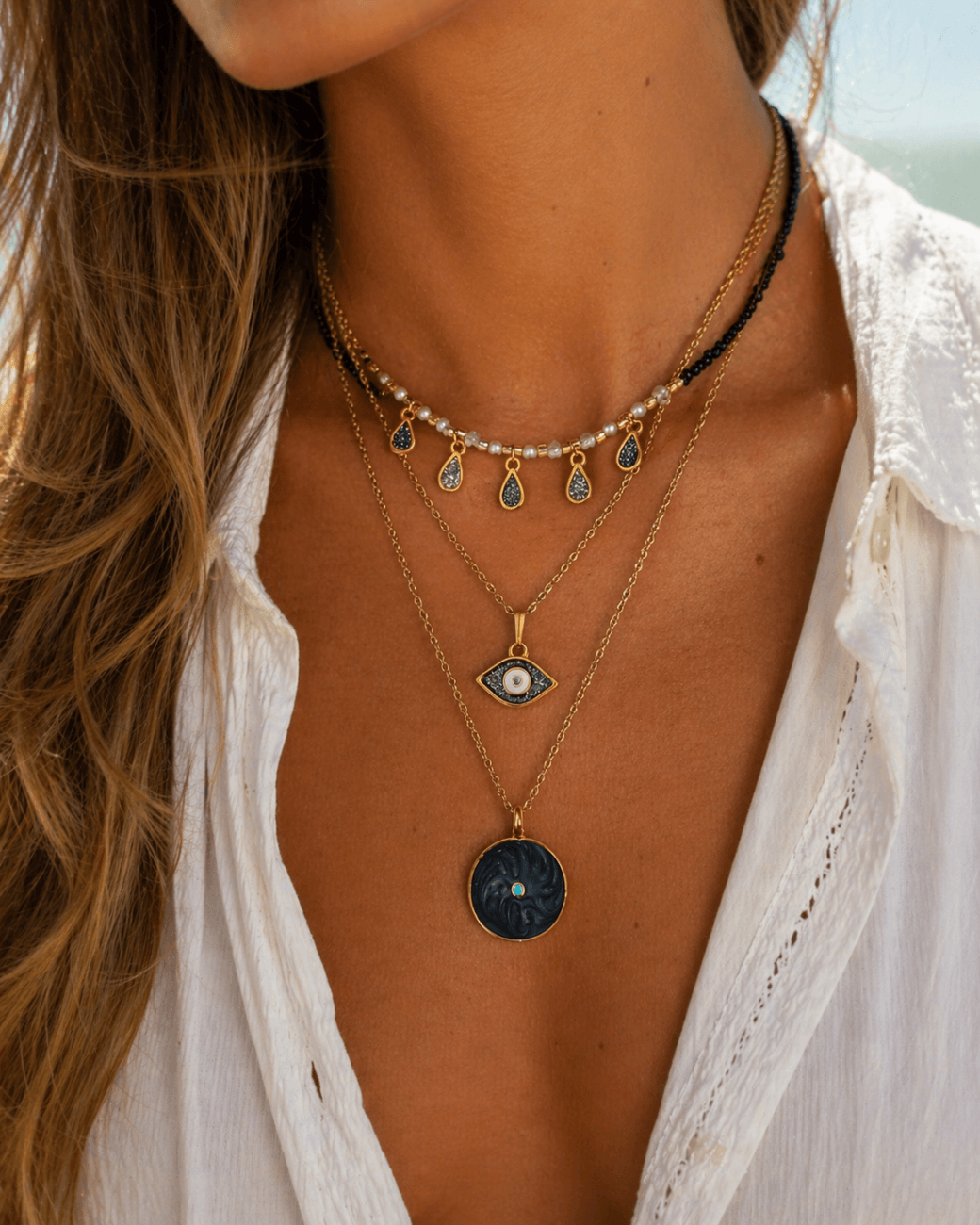 Τρία γυναικεία κολιέ layering με charms και μοτίφ μάτι φορεμένα μαζί σε λευκό πουκάμισο